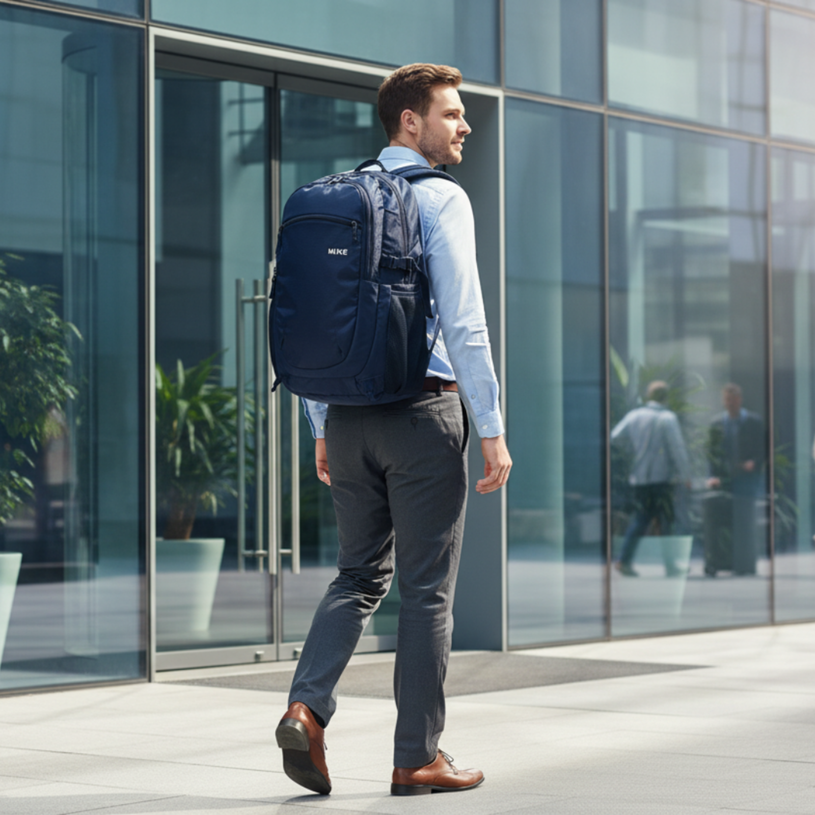 A man in a light blue shirt and gray trousers walks past a modern glass office building wearing a navy blue professional backpack. The backpack features a zippered main compartment, side pockets, and a small white logo on the upper right side.