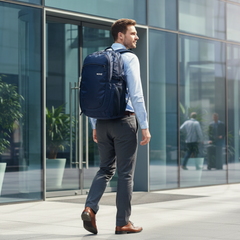 A man in a light blue shirt and gray trousers walks past a modern glass office building wearing a navy blue professional backpack. The backpack features a zippered main compartment, side pockets, and a small white logo on the upper right side.